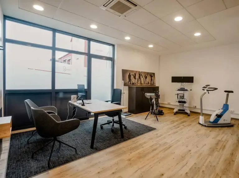 Moderner Behandlungsraum mit Holzboden und einem dunklen Teppich in einer Schräg-Ansicht. Im Vordergrund auf der linken Seite ist ein Tisch mit Stühlen zu sehen. Im Hintergrund auf der rechten Seite befinden sich verschiedene Geräte zur Ausdauermessung