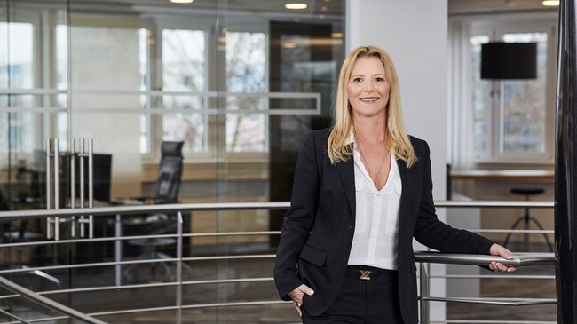 Isabella Leßwing – Mitglied im Führungsteam der offino Bürolösungen GmbH – posiert in dunkler Geschäftskleidung und sympathisch lächelnd vor dem Treppenabgang im ersten Stock des offino Showrooms in Kempten