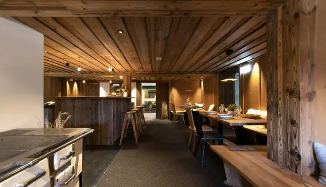 Hölzernes Hotel-Restaurant mit einer dunklen, aber gemütlichen Ästhetik. Auf der linken Seit ist eine Bar mit Barhockern zu sehen, auf der rechten Seite befinden sich gedeckte Tische mit Stühlen