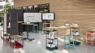 Arbeitsplatz vor einer hölzernen Wand, die aus einzelnen, aufeinander liegenden Balken besteht. Der Bereich ist durch ein Regal und einen hohen Tisch abgegrenzt und beinhaltt ein Whiteboard und einem portablen Bildschirm für Präsentationen.