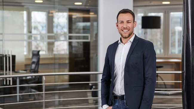 Fabio Leßwing – Mitglied im Führungsteam der offino Bürolösungen GmbH – posiert in dunkler Geschäftskleidung und sympathisch lächelnd vor dem Treppenabgang im ersten Stock des offino Showrooms in Kempten