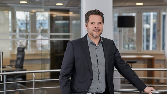 Jörg Wegmann – Mitglied im Führungsteam der offino Bürolösungen GmbH – posiert in dunkler Geschäftskleidung und sympathisch lächelnd vor dem Treppenabgang im ersten Stock des offino Showrooms in Kempten