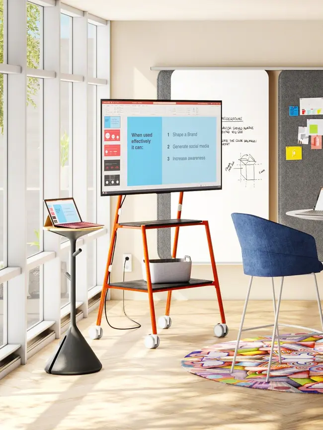 Aufstell- und fahrbarer Bildschirm, der eine Powerpoint-Präsentation darstellt und sich in einem gemütlichen Arbeitsraum befindet. Neben dem Bildschirm ist ein modernes Posest mit einem Tablet mit Tastatur platziert, auf dem die selbe Präsentationsdarstellung erfolgt.