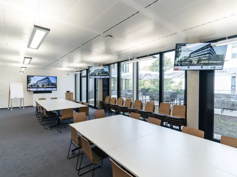 Meeting Room im Showroom der Experten für nachhaltige Raumgestaltung. Zwei große weiße Tische, umgeben von gleichmäßig platzierten Holzstühlen. An den Wänden hängen insgesamt drei Bildschirme, die sich zum Darstellen einer Präsentation eignen. An einer Fensterwand im Hintergrund befinden sich weitere, aufeinander gestapelte Stühle.