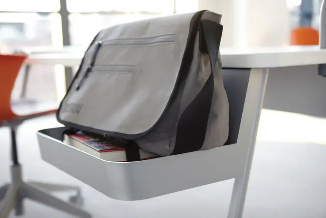 Nahaufnahme einer integrierten Seitenablage eines Unterrichtstisches zum Ablegen von Taschen und Büchern. In der Ablage befindet sich eine graue Umhängetasche