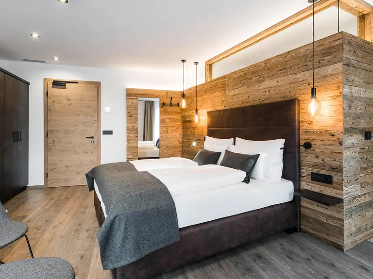 Modernes Hotelzimmer im rustikal-traditionellen Industrial-Style: ein Boxspringbett mit dunklem Lederbezug, dunkel marmoriertem Kleiderschrank, und einem Raumteiler aus Echtholz zum modernen Badezimmer mit freistehender Badewanne hin – ein Beispiel der Arbeit der offino Bürolösungen GmbH im Bereich RÄUME & WOHLFÜHLEN.