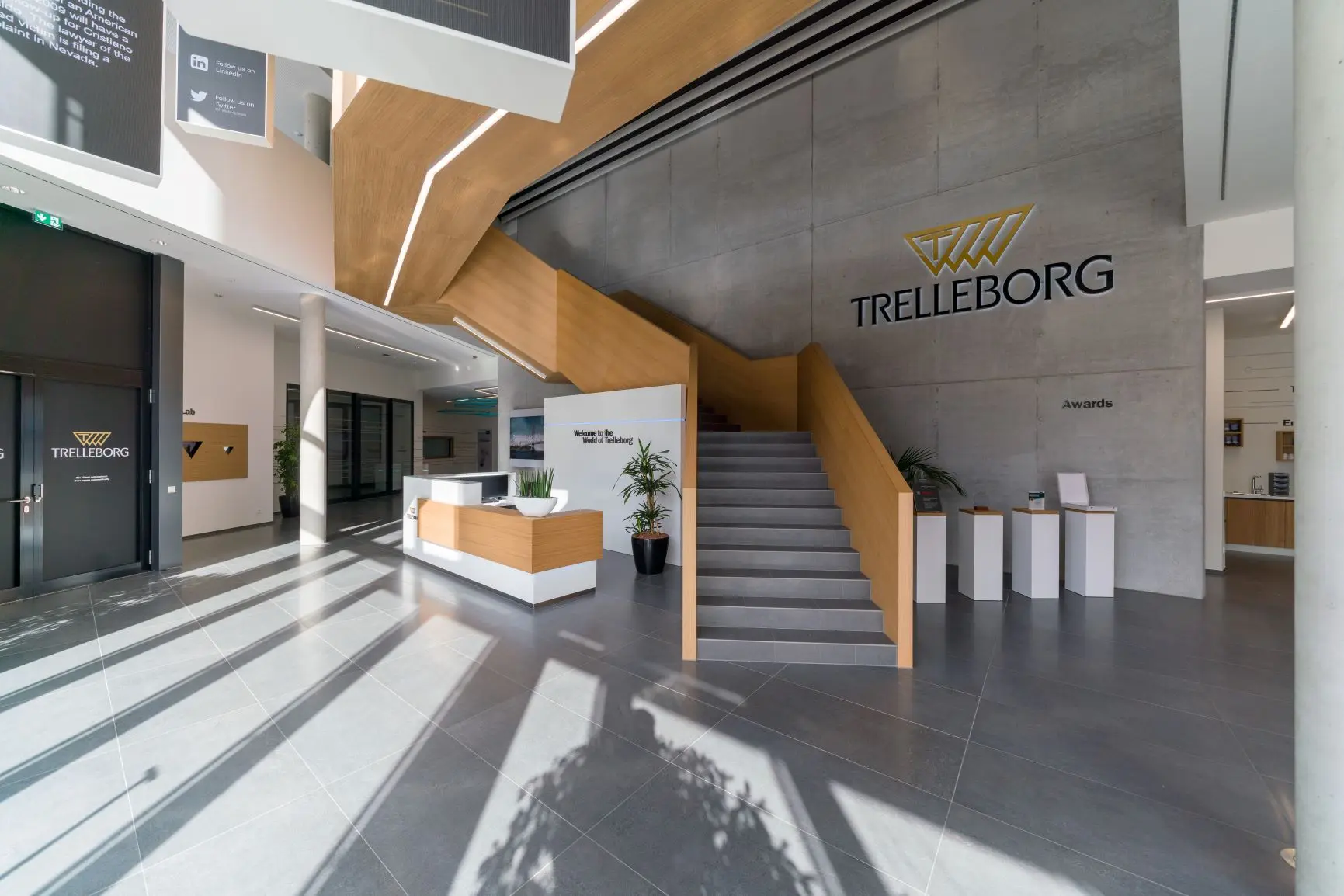 Die Rezeption im offino Showroom, Ihre Experten für nachhaltige Büro- und Raumgestaltung. Ein großer Saal, an dem eine holzverkleidete Rezeptionstheke steht. Rechts neben ihr sieht man einen Treppenaufgang, der in ein höheres Stockwerk führt.