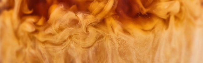 Detailansicht im Querschnitt: Milschaum oder Milch wird in Kaffee gegeben und mischt sich mit selbigem. Besonders an der Grenzen zwischen den beiden Flüssigkeiten bildet der Milchschaum dabei dekorative und verspielte Muster. Das Bild repräsentiert den Fachbereich KAFFEE & GENUSS der offino Bürolösungen GmbH.