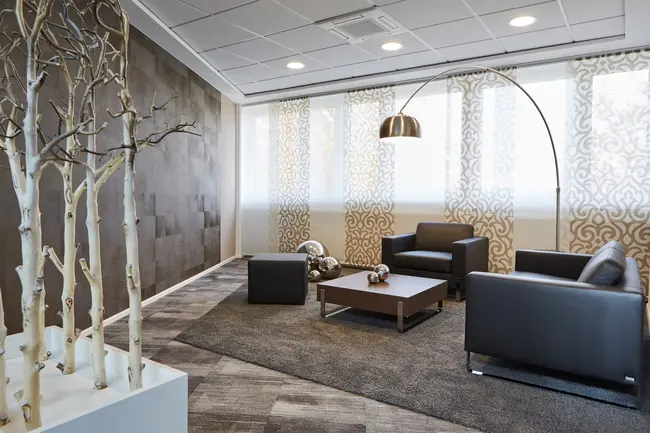 Gemütliche Möbelgruppe für Besprechungen in einem in braun gehaltenen Raum des Showrooms der offino Bürolösungen GmbH in Kempten. Ein Hocker, Ein Sofa, ein Sessel und eine gebogende Stehlampe werden von einem Kunstobjekt aus Birkenstämmen und einem weißen Sockel am linken Bildrand in Szene gesetzt.