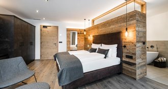 Hotelraum mit Holzboden und Holzwänden. Im Fokus liegt ein Doppelbett, das an der hölzernen Wand platziert und von modernen Lampen umgeben ist.