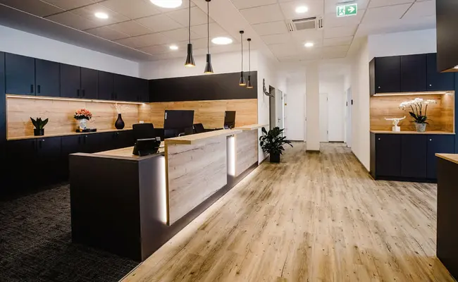 Schräg-Ansicht einer ästhetischen Theke als Rezeption von modernen Behandlungsräumen. Hinter der Theke sind zwei Bildschirme und ein dunkles, hölzernes Regal zu sehen. Auf der rechten Seite sieht man in einen Gang hinein, in dessen Mitte sich eine Säule befindet.