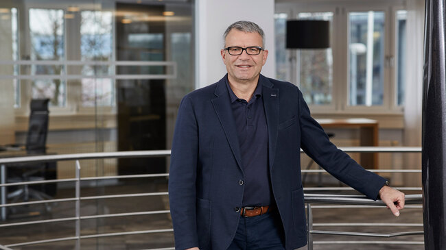 Horst Leßwing – Mitglied im Führungsteam der offino Bürolösungen GmbH – posiert in dunkler Geschäftskleidung und sympathisch lächelnd vor dem Treppenabgang im ersten Stock des offino Showrooms in Kempten