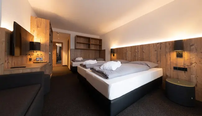 Hotelzimmer mit hölzernem Ambiente. Zu sehen ist ein gemütliches Doppelbett, dahinter befindet sich ein gleich aussehendes Einzelbett.