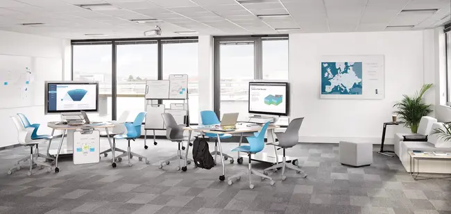 Große Ansicht eines Unterrichts- und Lehrraums mit runden Gruppentischen mit verschiedenfarbigen Bürostühlen. An der Wand im Hintergrund ist ein Bildschirm für Präsentationen und Infografiken platziert.